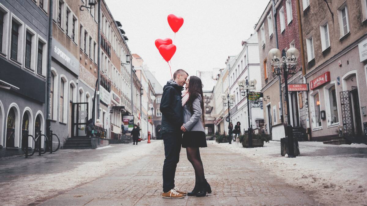 A couple standing in the middle of a slow city street, very close to one another, smiling, th eman holding three red heart-shaped balloons.