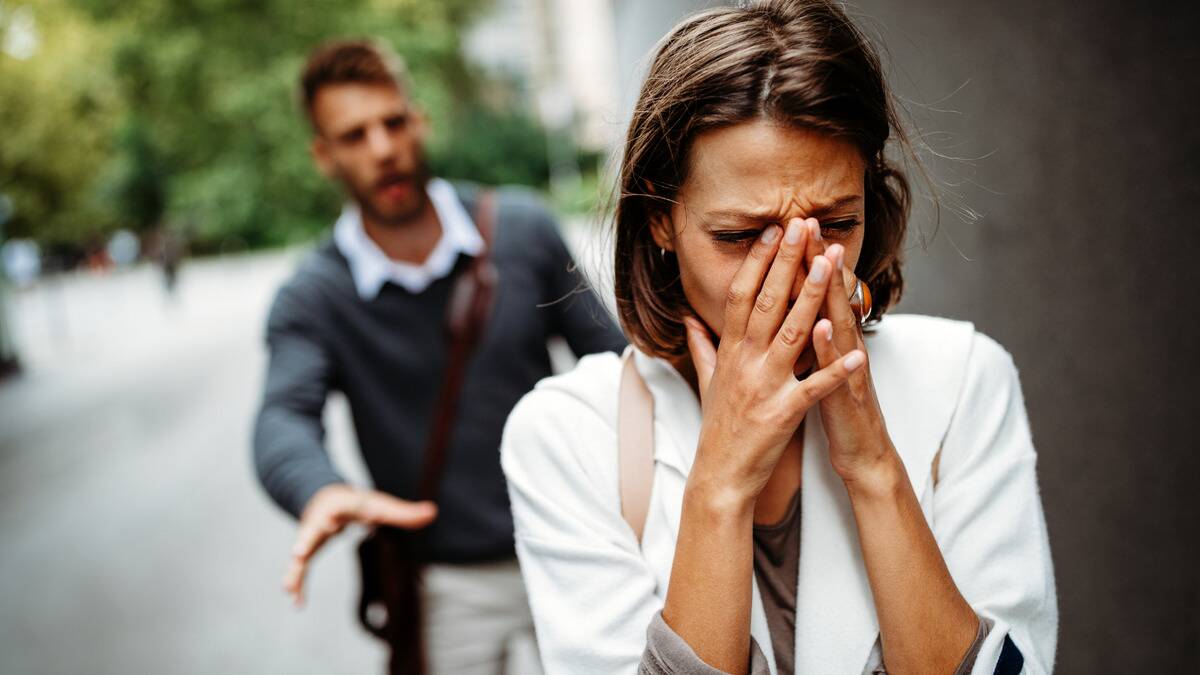 A woman walking away from a man crying, the man seen in the background reaching out to try and stop her.