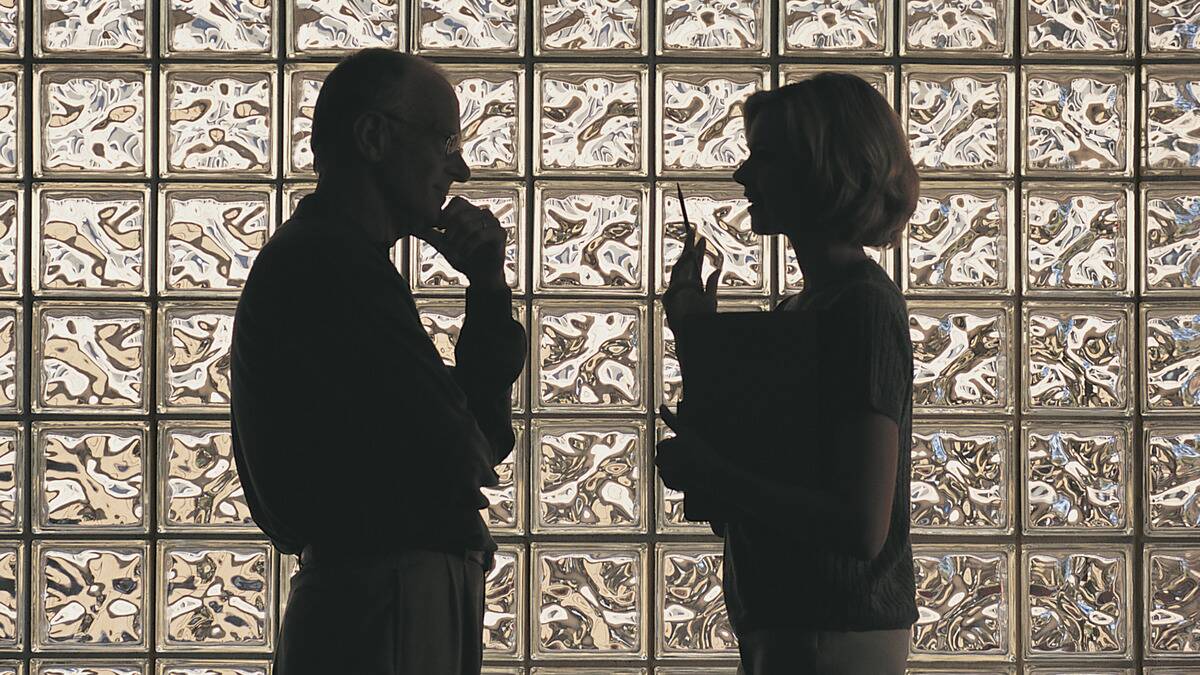 A man and woman talking, standing a few feet apart, silhouetted by the glass wall behind them.