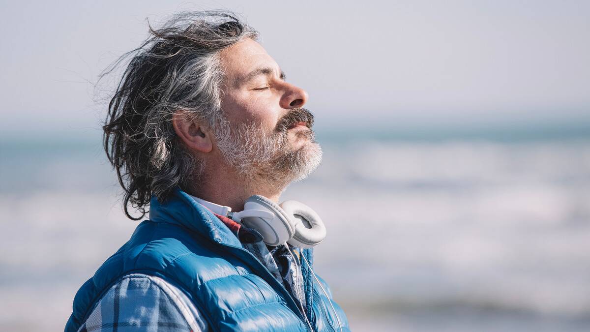 A man standing on the beach by the water, eyes closed, head tilted back as the wind blows his hair back.