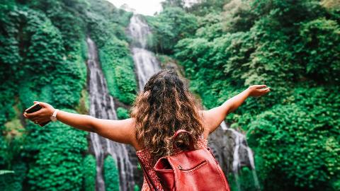 Someone photographed from behind, her arms spread out in awe as she looks at a beautiful waterfall surrounded by greenery.