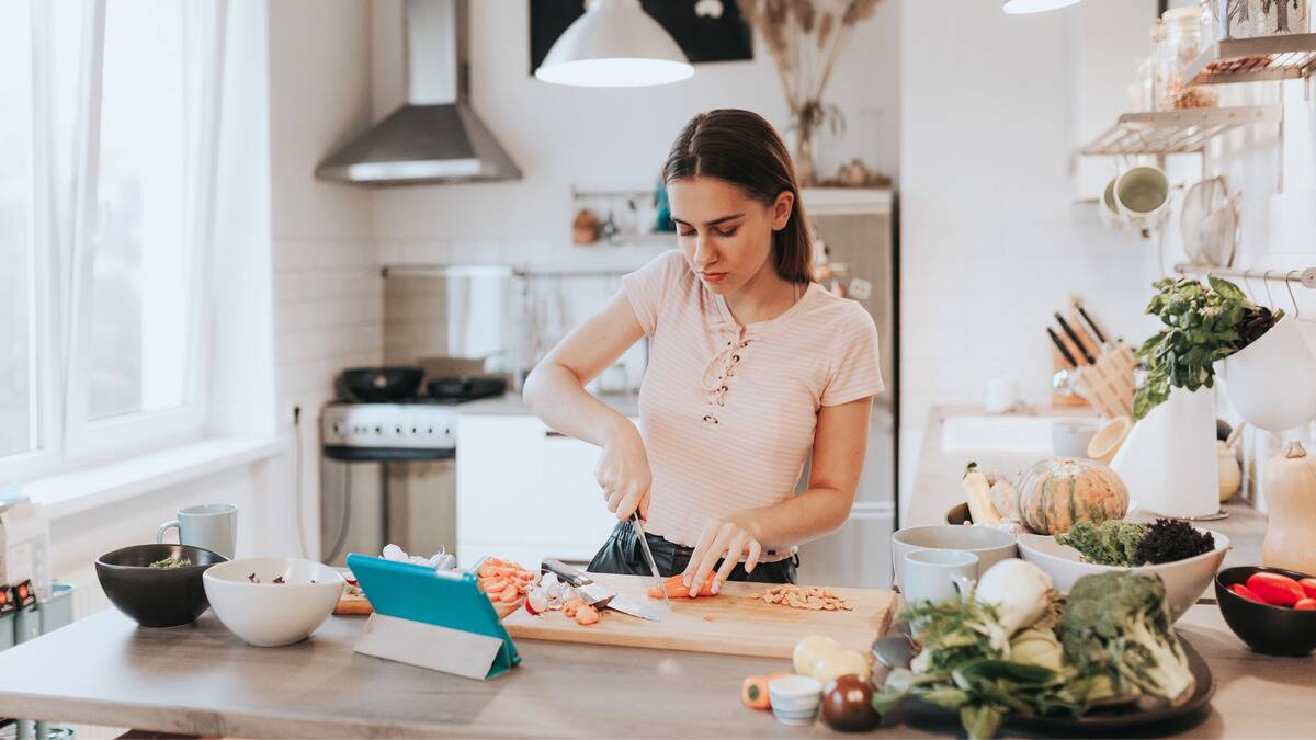 A woman learning how to cook, chopping veggies while watching a tutorial on her tablet.