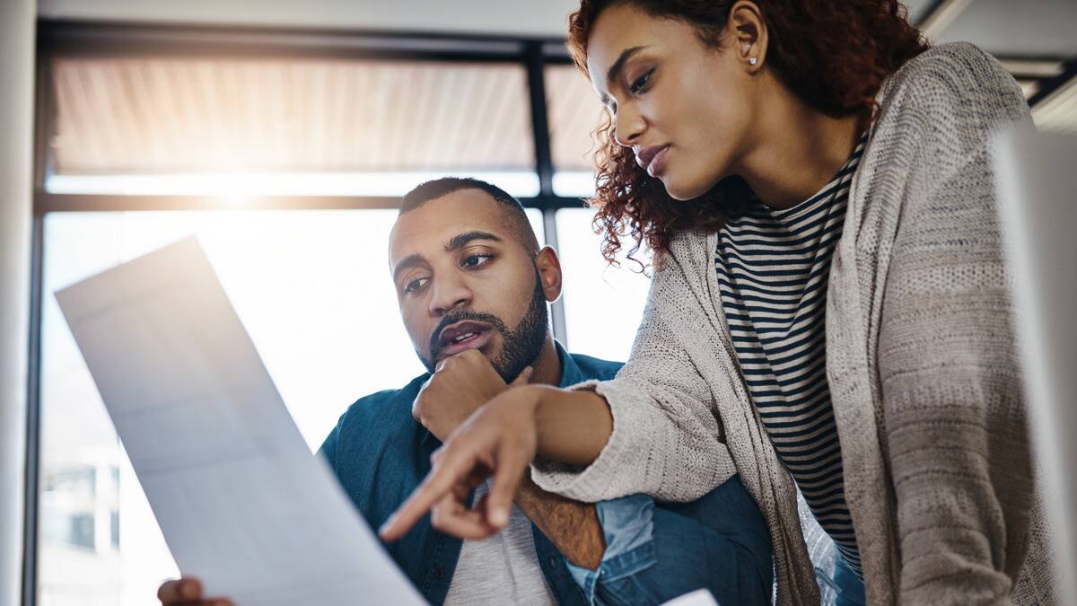 A couple both reviewing financial documents, the woman standing and pointing something out while the man is sitting with his hand on his chin.