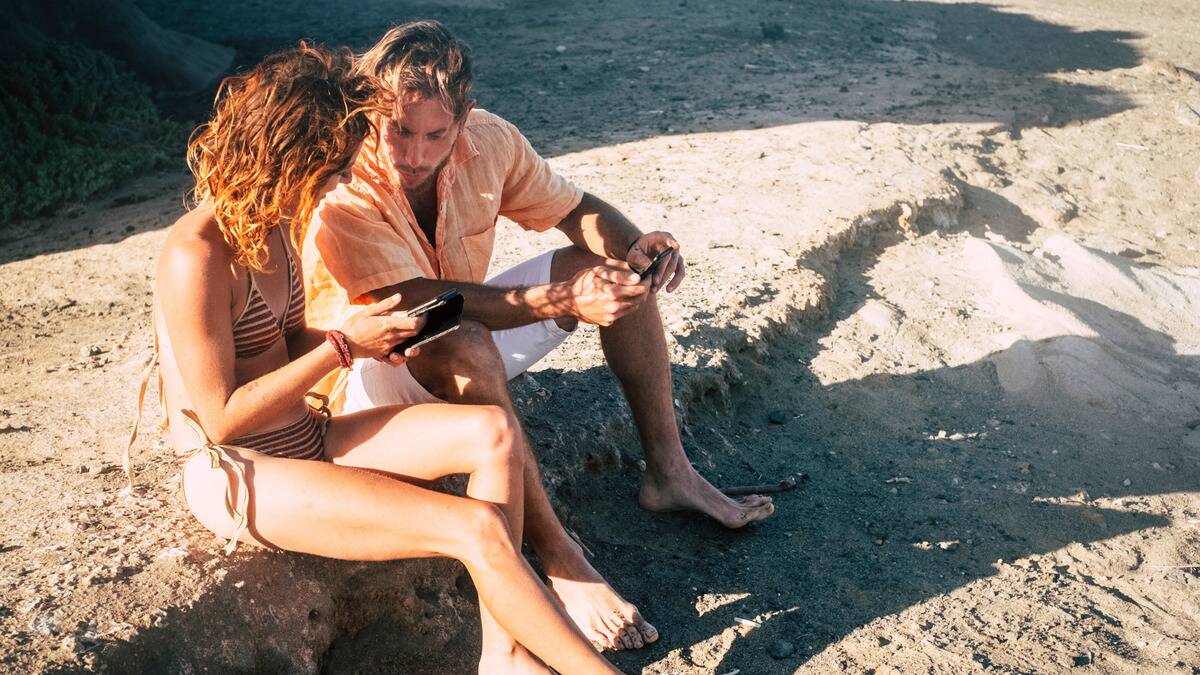 A couple sitting on the beach, the woman showing the man something on her phone.