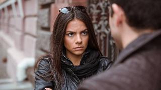 A woman looking very sternly at a man she's talking to as they stand outside on a city street.
