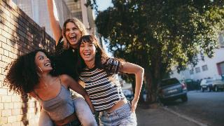 Three friends walking through the street together, one giving another a piggy back ride, all laughing and smiling.