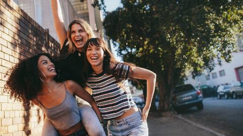Three friends walking through the street together, one giving another a piggy back ride, all laughing and smiling.