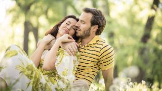 A couple sitting in the grass together, the woman leaning into the man's chest, the man's arm around her.