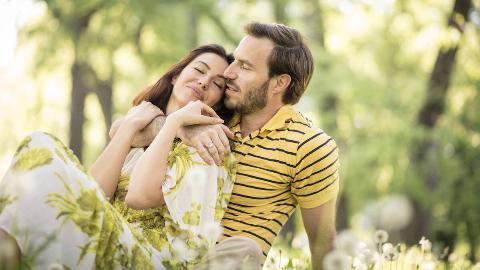 A couple sitting in the grass together, the woman leaning into the man's chest, the man's arm around her.
