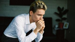 A man sitting in an office, leaning forward, chin in his hands, looking very pensive.