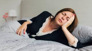 A woman leaning her head in her hand as she lays on her side in bed, eyes closed, looking tired.