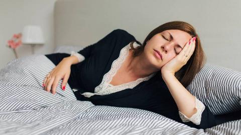 A woman leaning her head in her hand as she lays on her side in bed, eyes closed, looking tired.