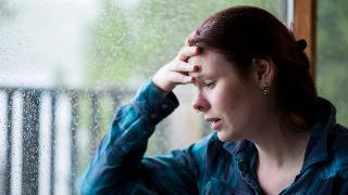 A woman sitting by a window as it rains outside, a hand on her forehead, looking as if she's been crying.