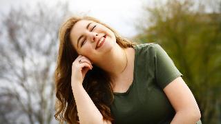 A woman smiling with her eyes closed as she stands outside, head tilted and leaning against her hand.