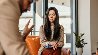 A woman sitting on a couch, legs and arms folded, looking insecure as a man speaks to her.