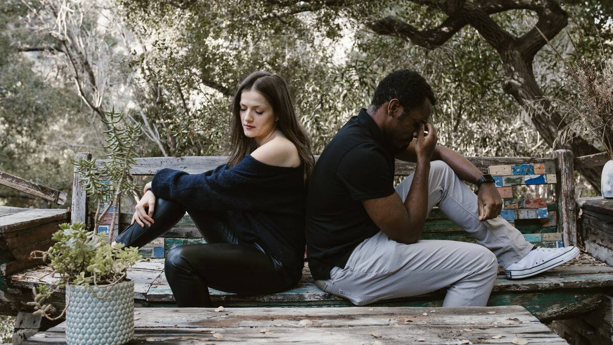 A man and woman sitting back to back on a bench, both looking sorrowful.