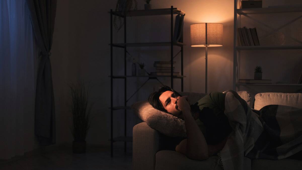 A dark photo of a man laying on his couch, a light highlighting his face, wher ehe's biting his thumbnail as he lays there stressed out.