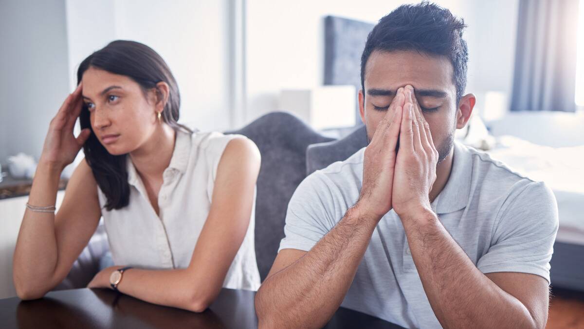 A man and woman sitting side by side at a table, the man covering his face with both hands, the woman with her fingers on her temple, looking annoyed.