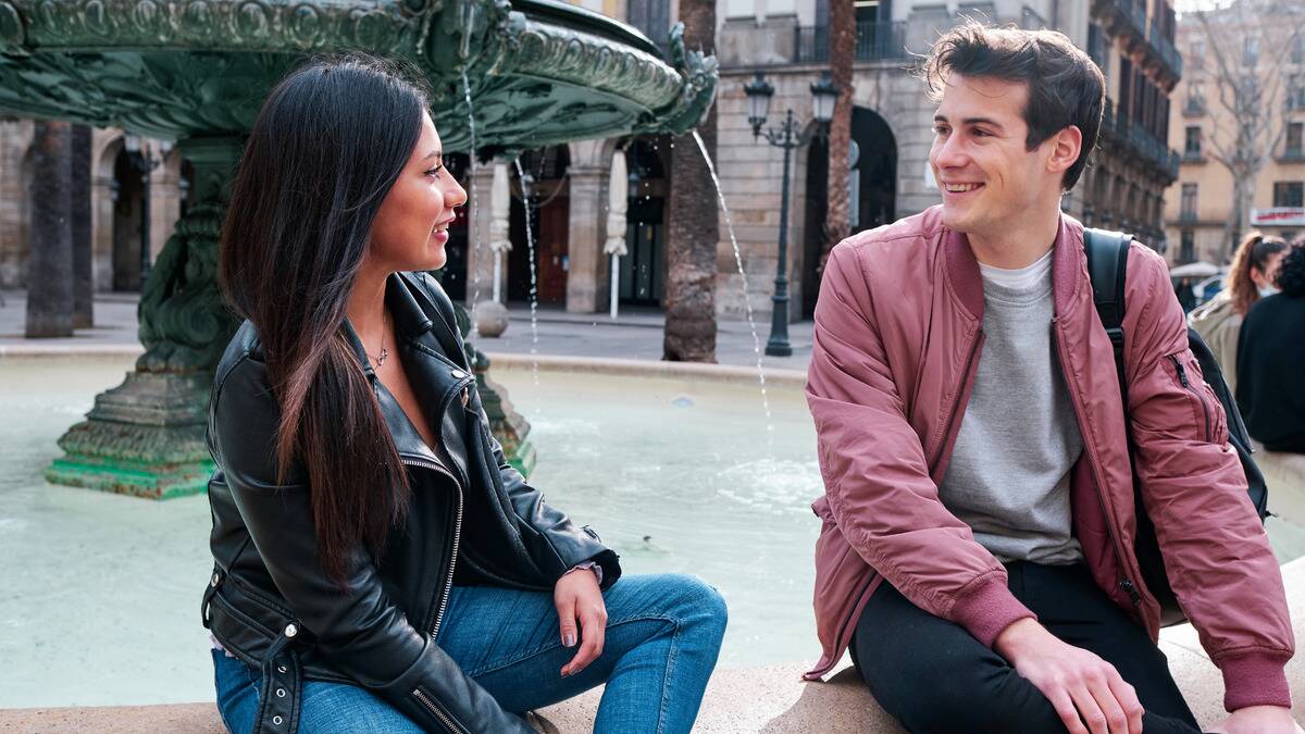 Two friends, a man and a woman, sitting next to each other by a fountain and chatting.