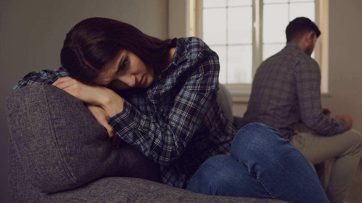 A woman curled up on the couch, looking sad as her boyfriend in the background ignores her.