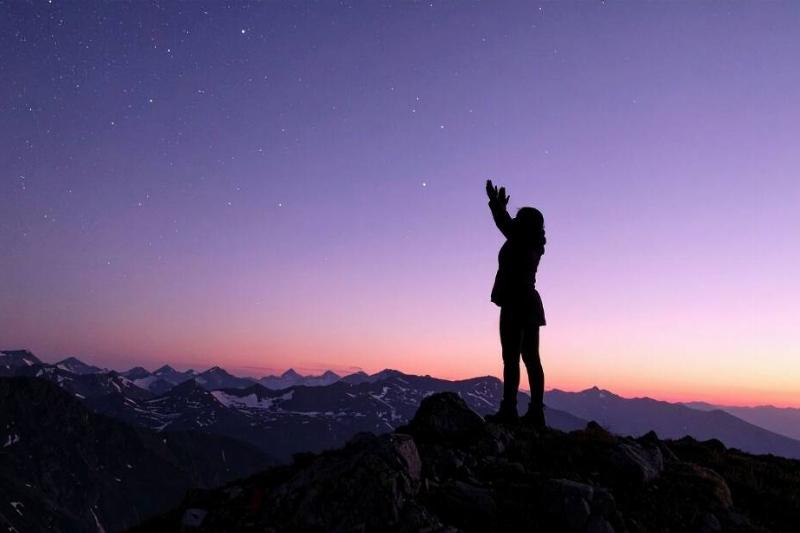 A woman standing on the top of a mountain, silhouetted, hands up to the stars