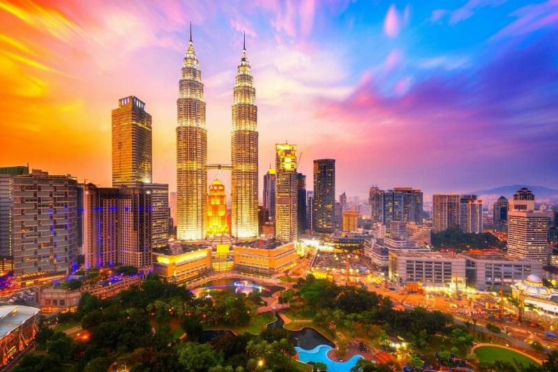 A beautiful photo of a city from Malaysia, with vibrantly-hued skies behind a cityscape, highlighting some unique interconnected towers. At the bottom of the image is a green area, presumably a park, with elegant water features