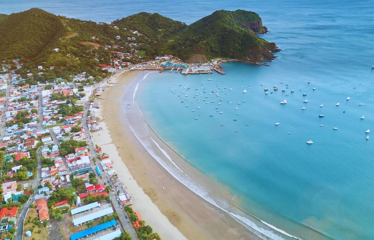 A shot of a shoreline in Nicaragua, showcasing elements of a charming city, natural beauty with some mountains, and a series of boats deeper in the water.