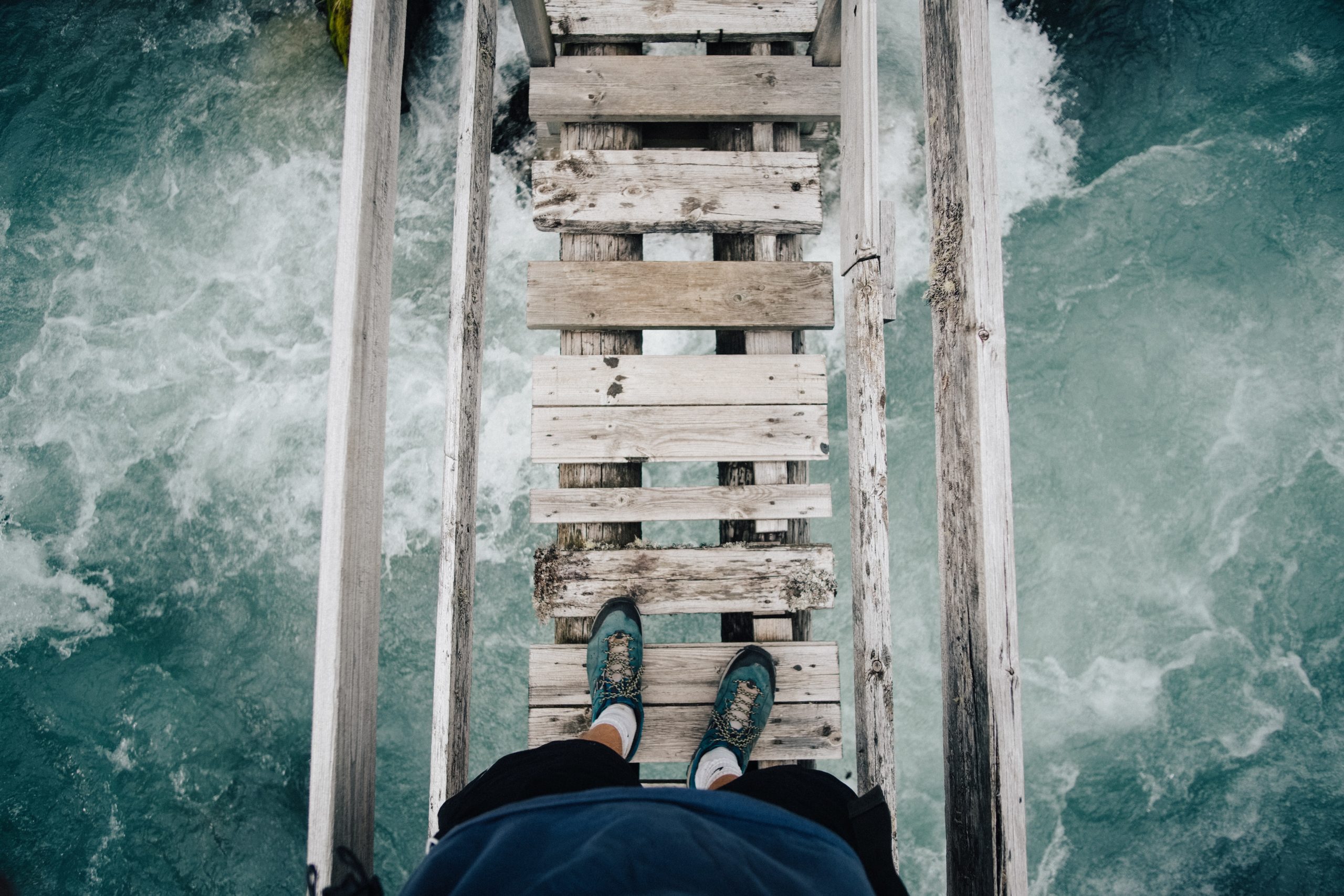 Someone standing on a rickety bridge above turbulent water.