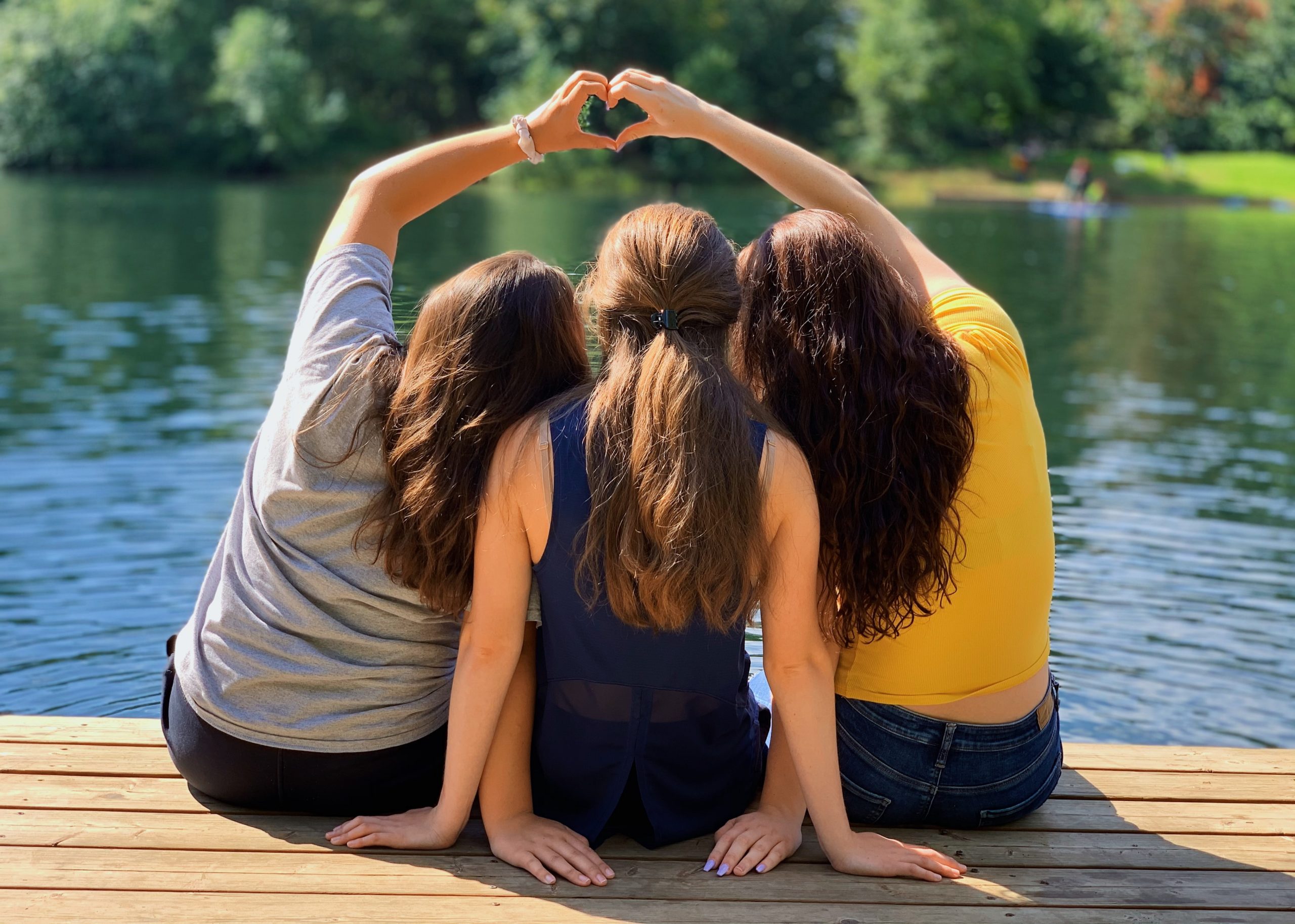 Three girls sitting on a dock, the two on the end making a heart with their head overtop.