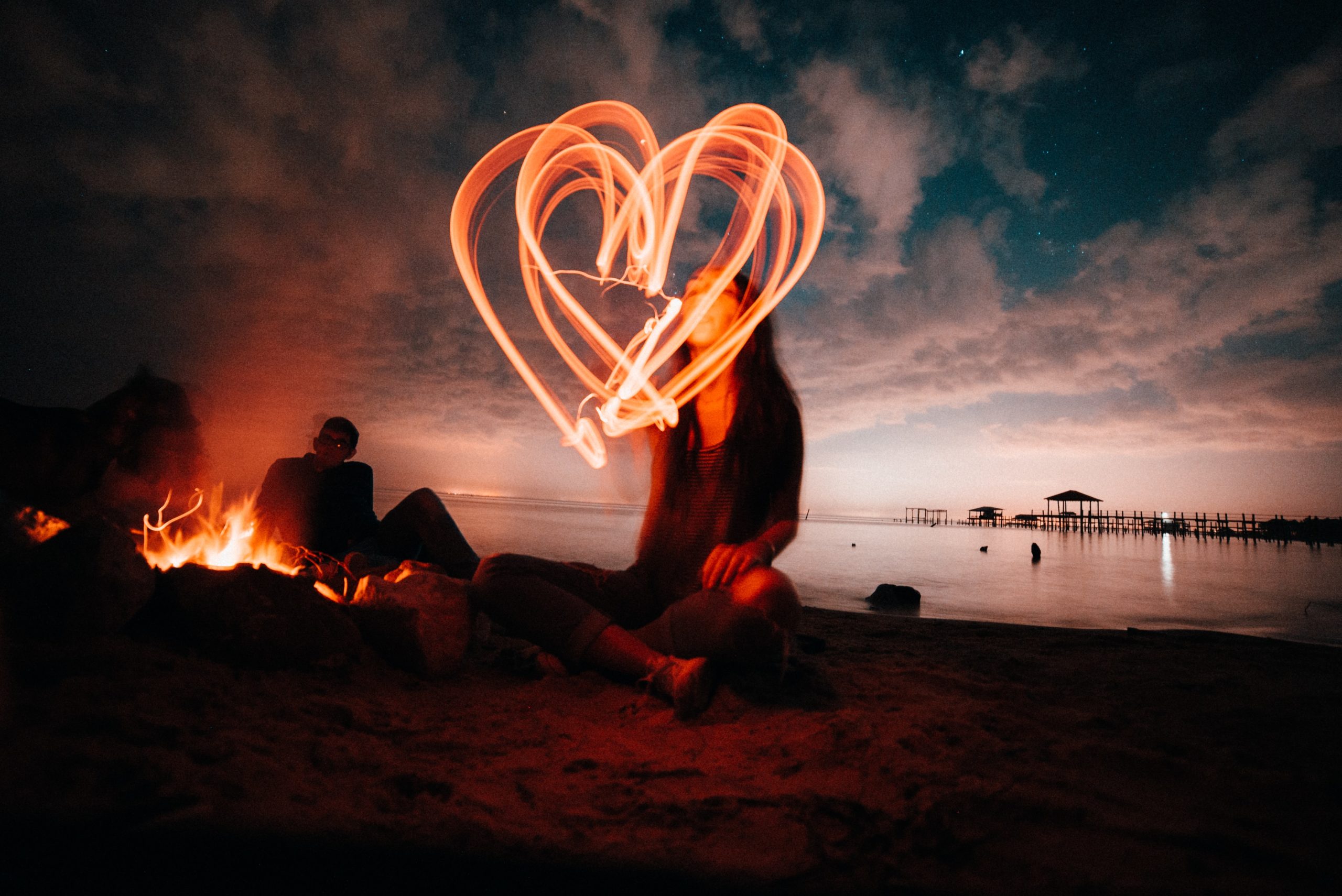 A long-exposure photo of a woman drawing a heart with a light source while seated on the beach.
