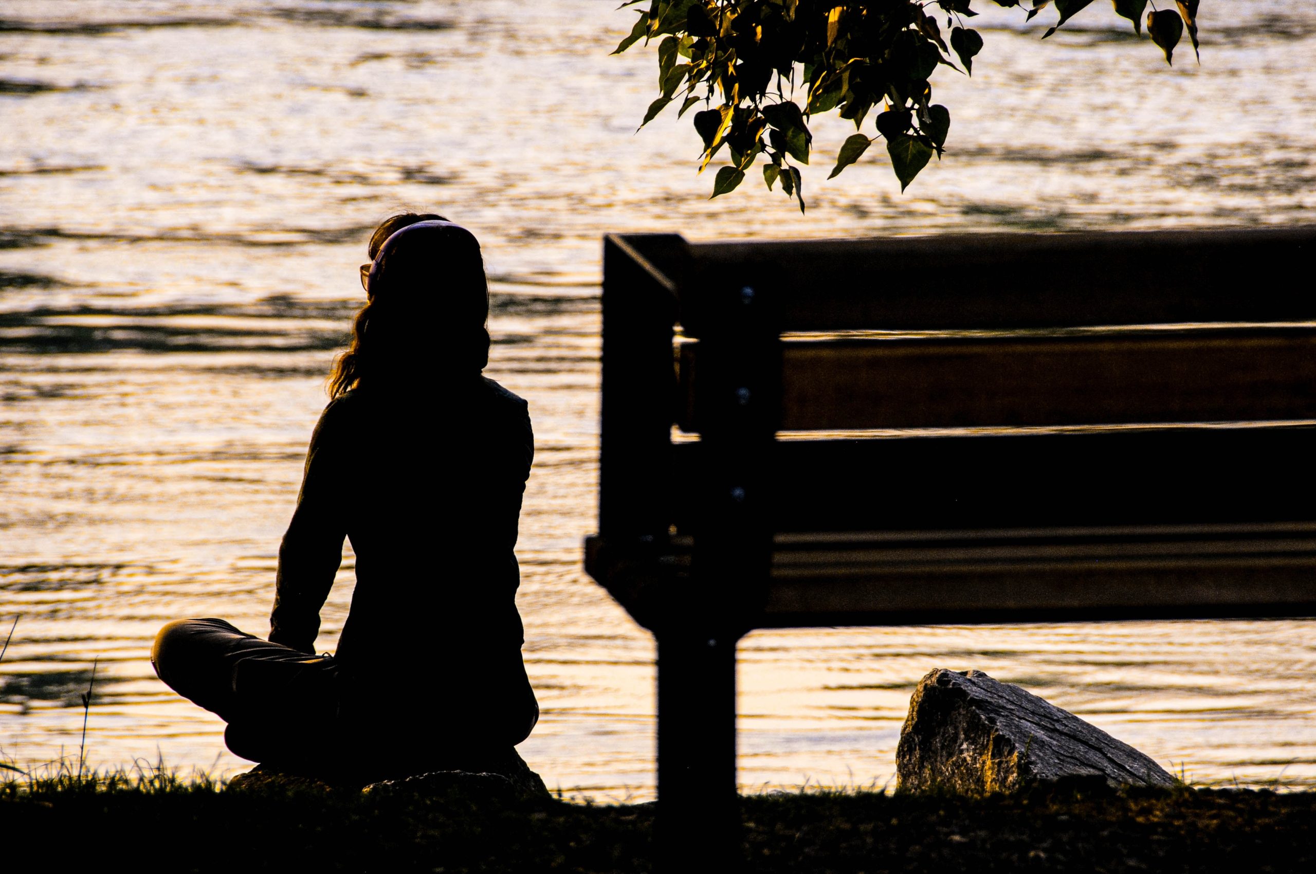 The silhouette of a woman meditating next to a park bench by water.