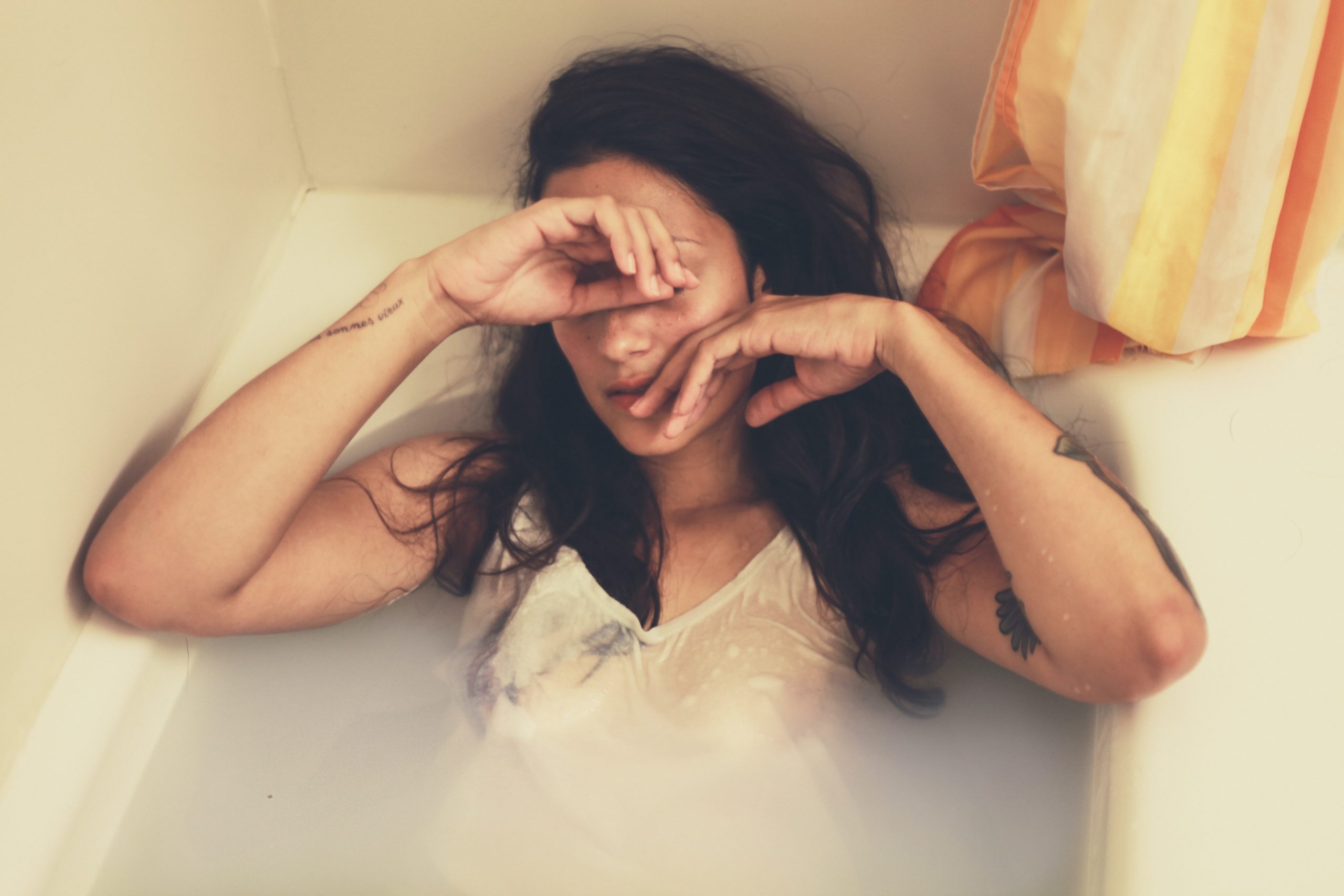 A woman laying in her bathtub with her shirt still on, hands covering her face.