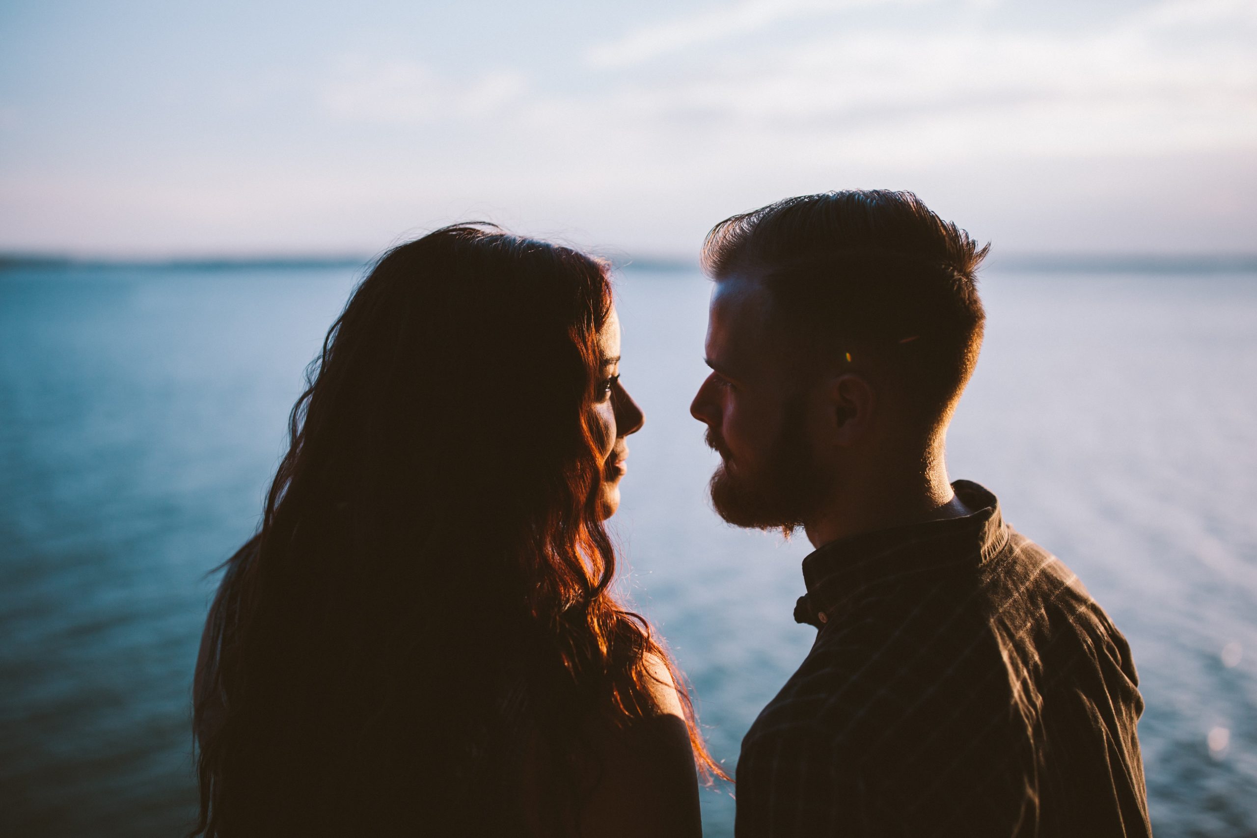 A couple smiling at one another as they stand before a lake.