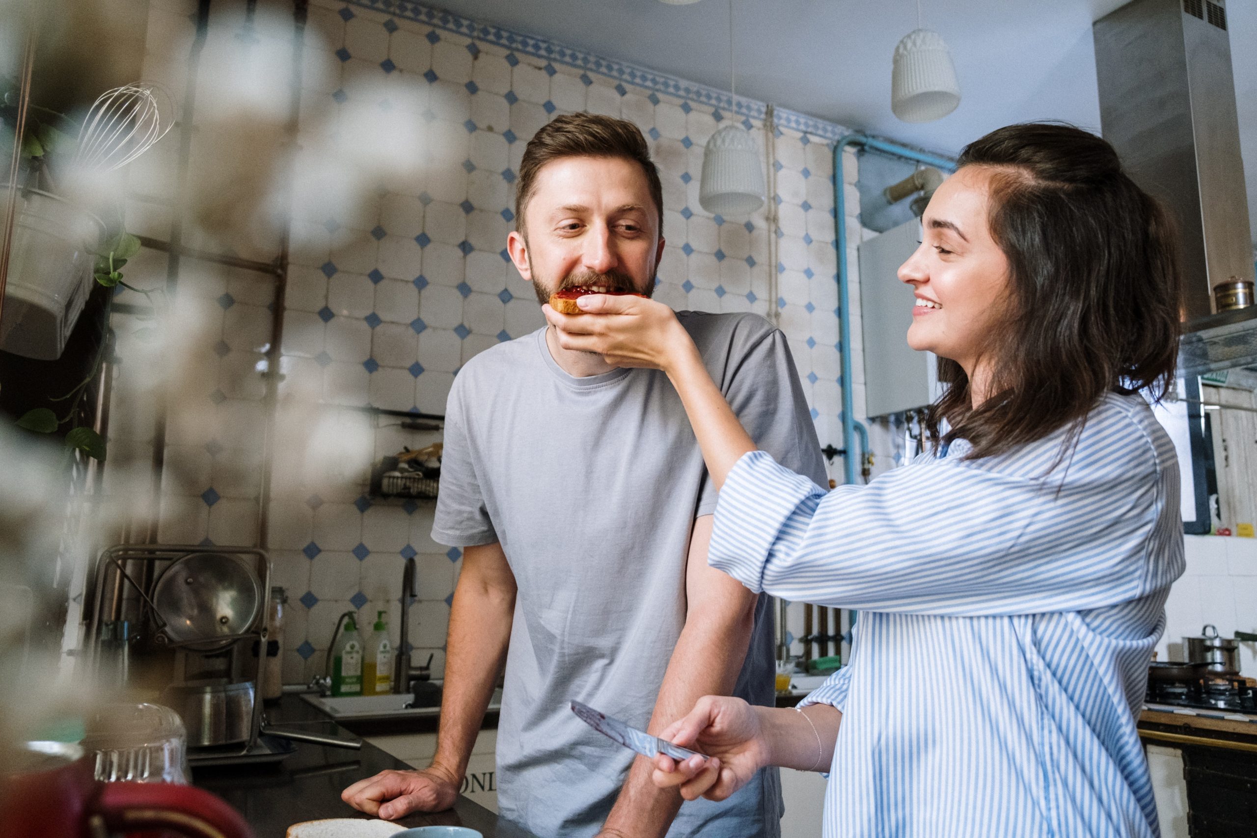 A couple in the kitchen, a woman feeding a man a bite of the food she's making.