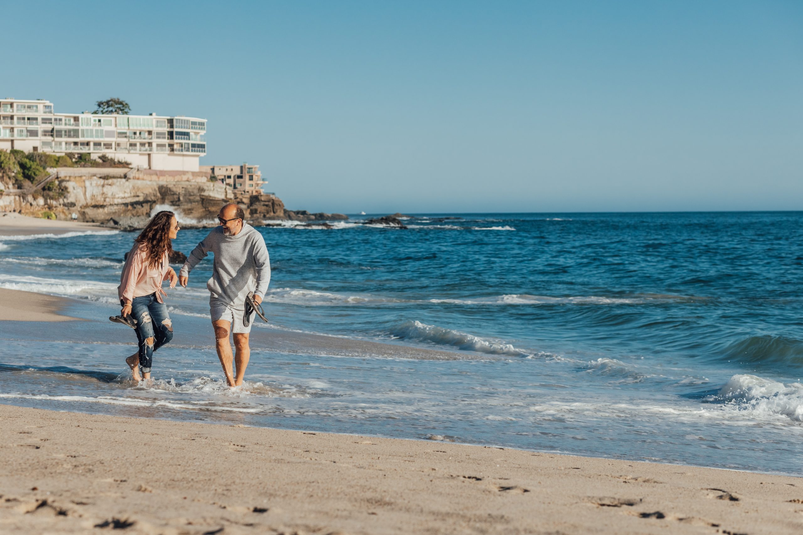 A couple walking along a beach, shoes off as they treat shallow water.