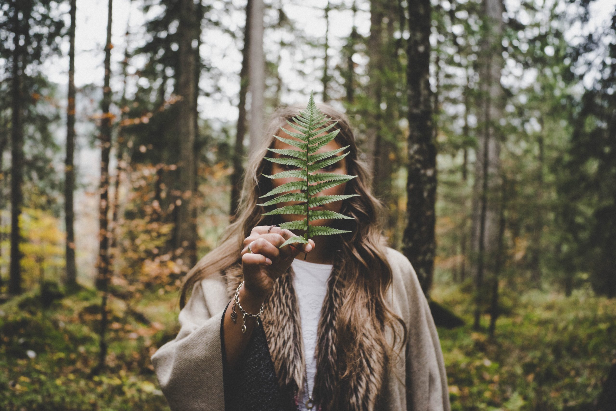 A woman standign in a forest, holding a leaf in front of her face.