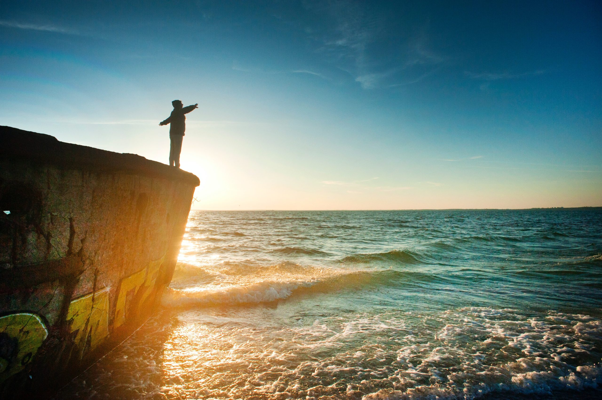 Someone standing on a cliff with their arms out, up above the water.