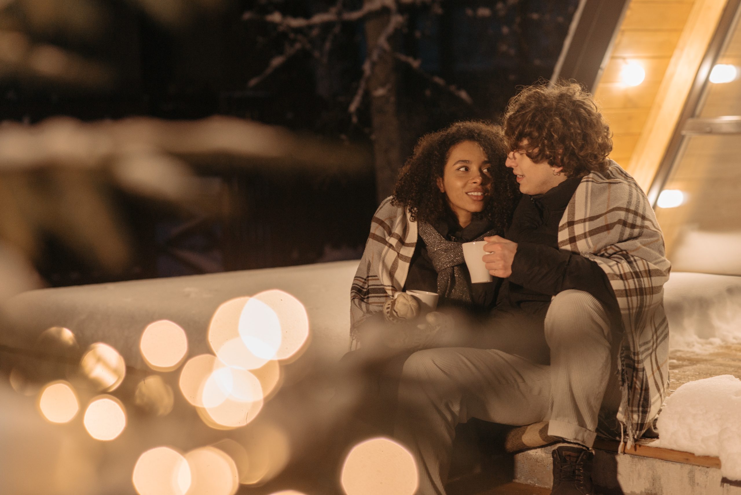 A couple sitting outside under a blanket, small lights scattered around them.