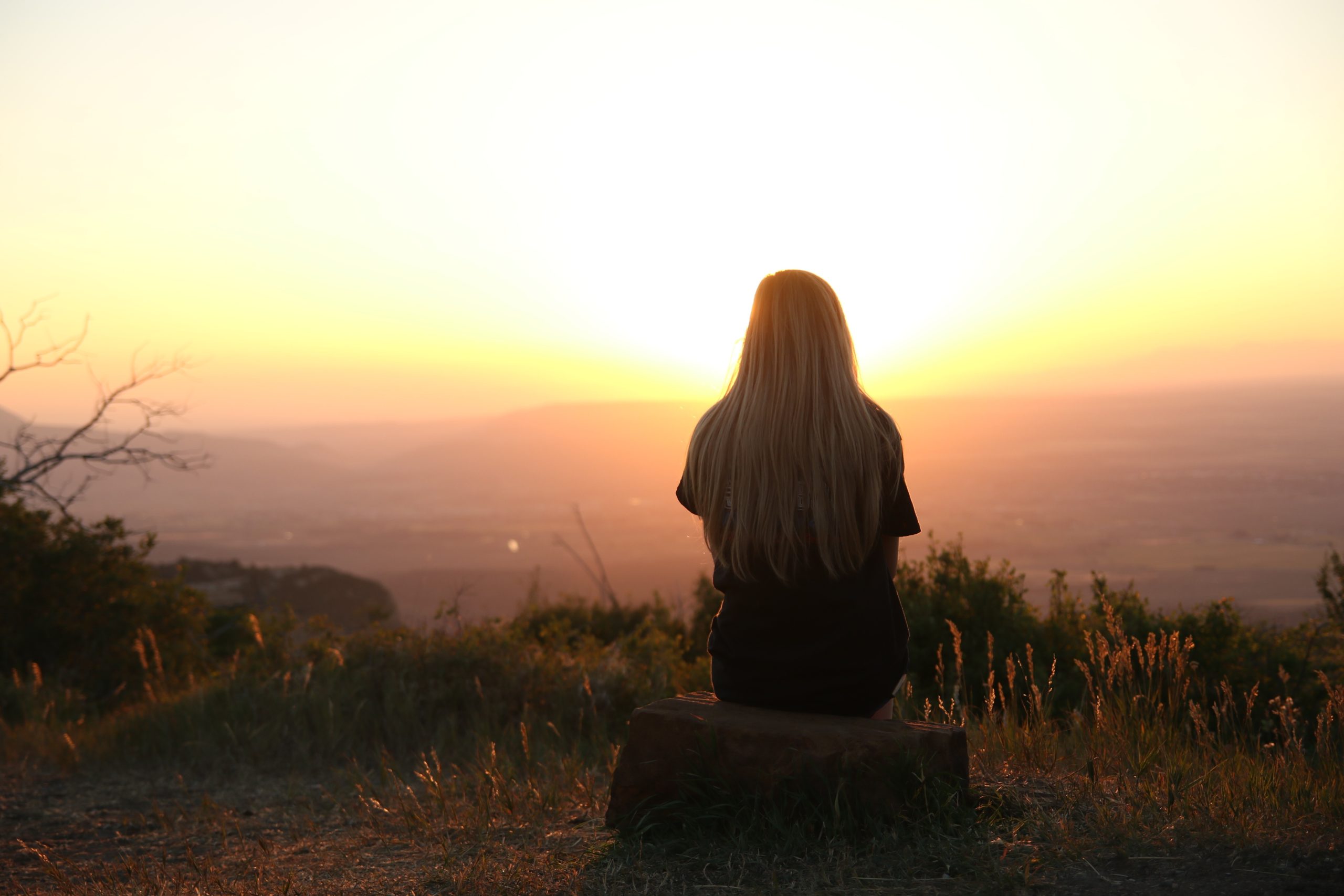 A woman sitting on a rock, facing a sunrise.