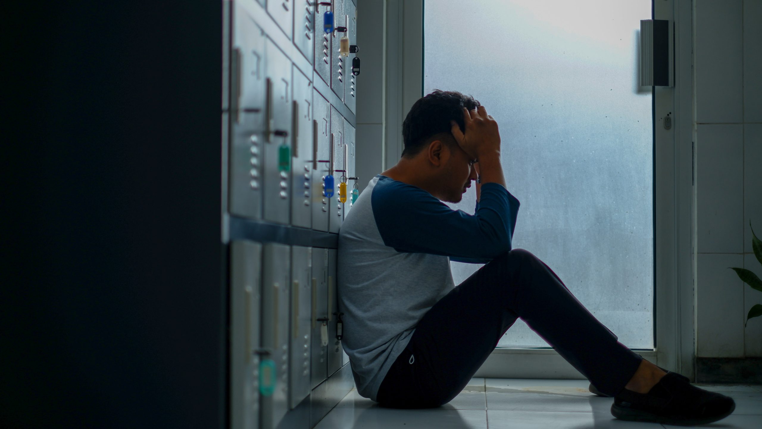 A man seated on the floor, leaning against some lockers, holding his own head.