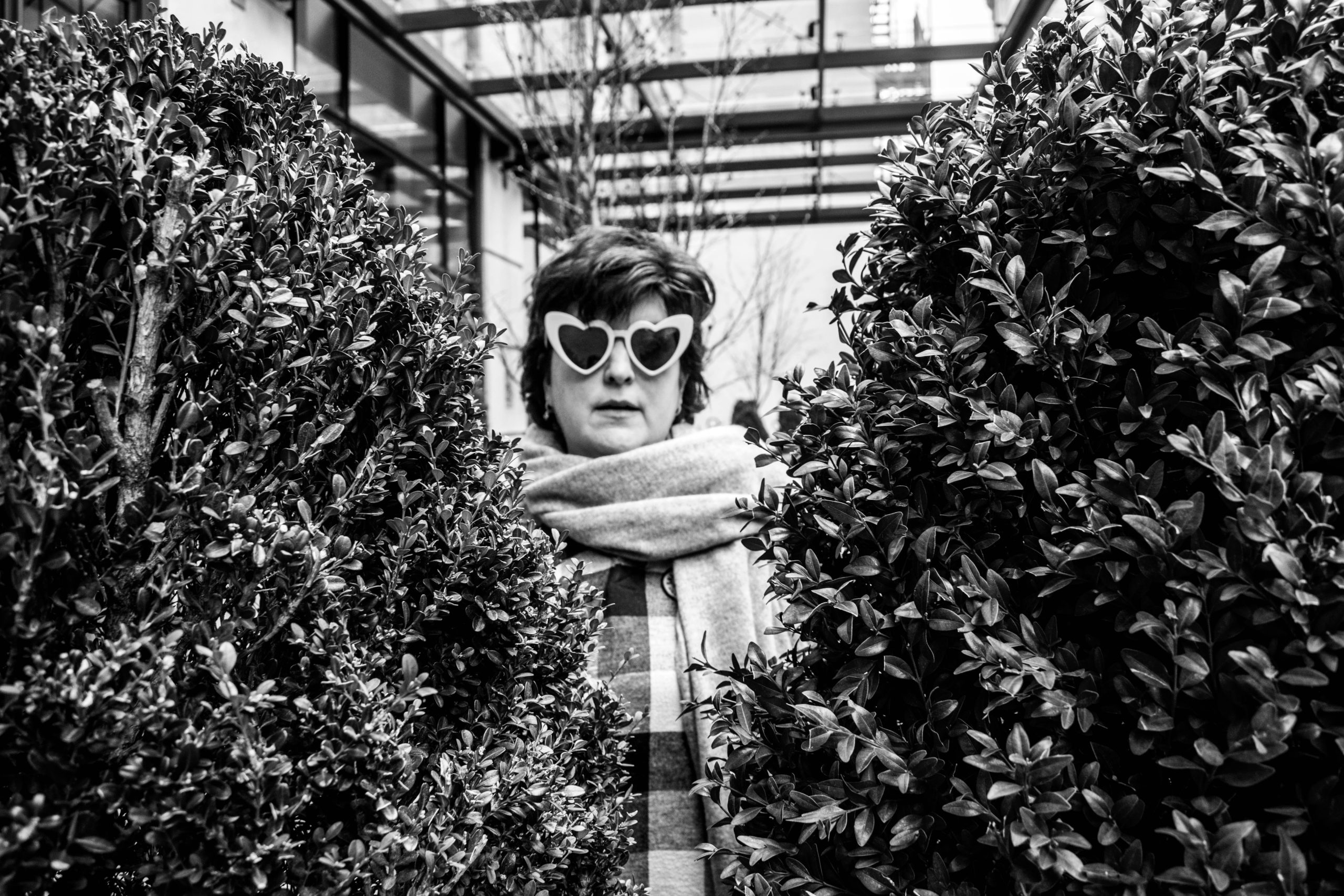 A greyscale image of a woman in heart glasses between some bushes.