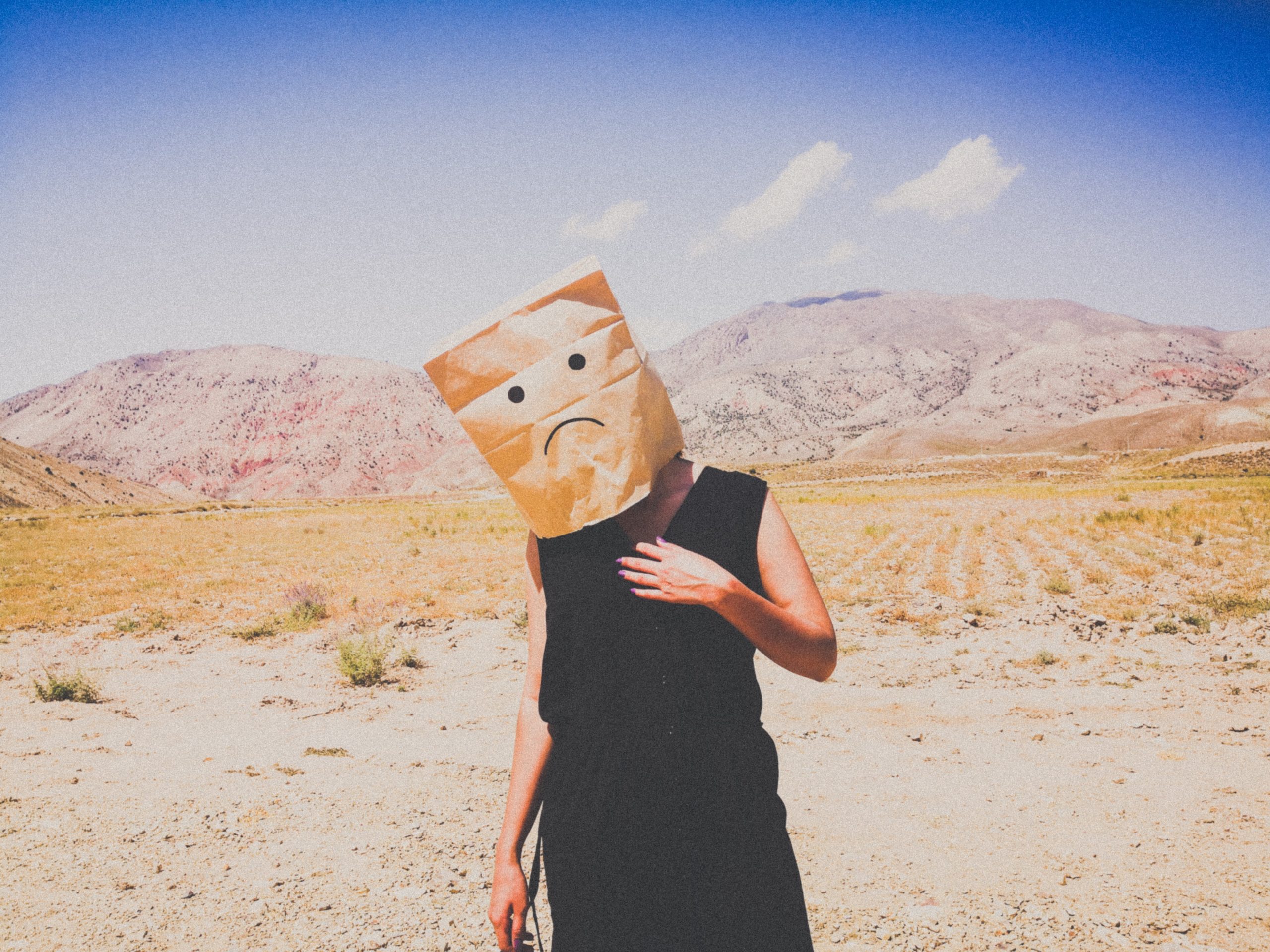 A woman standing in a desert plain with a bag over her head, a sad face drawn on it.