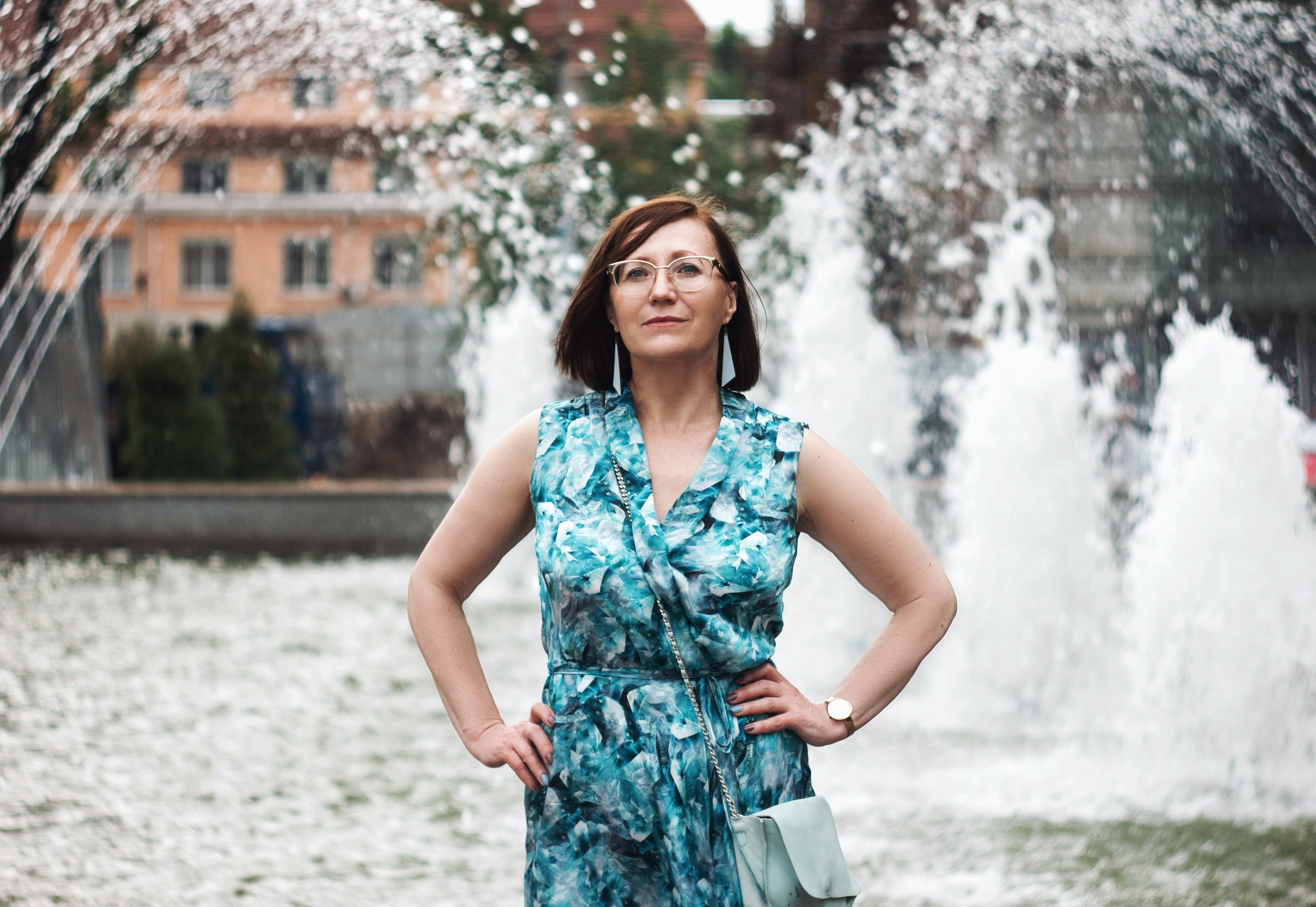 A woman standing in front of a fountain, hands on her hips, facing the camera head on.
