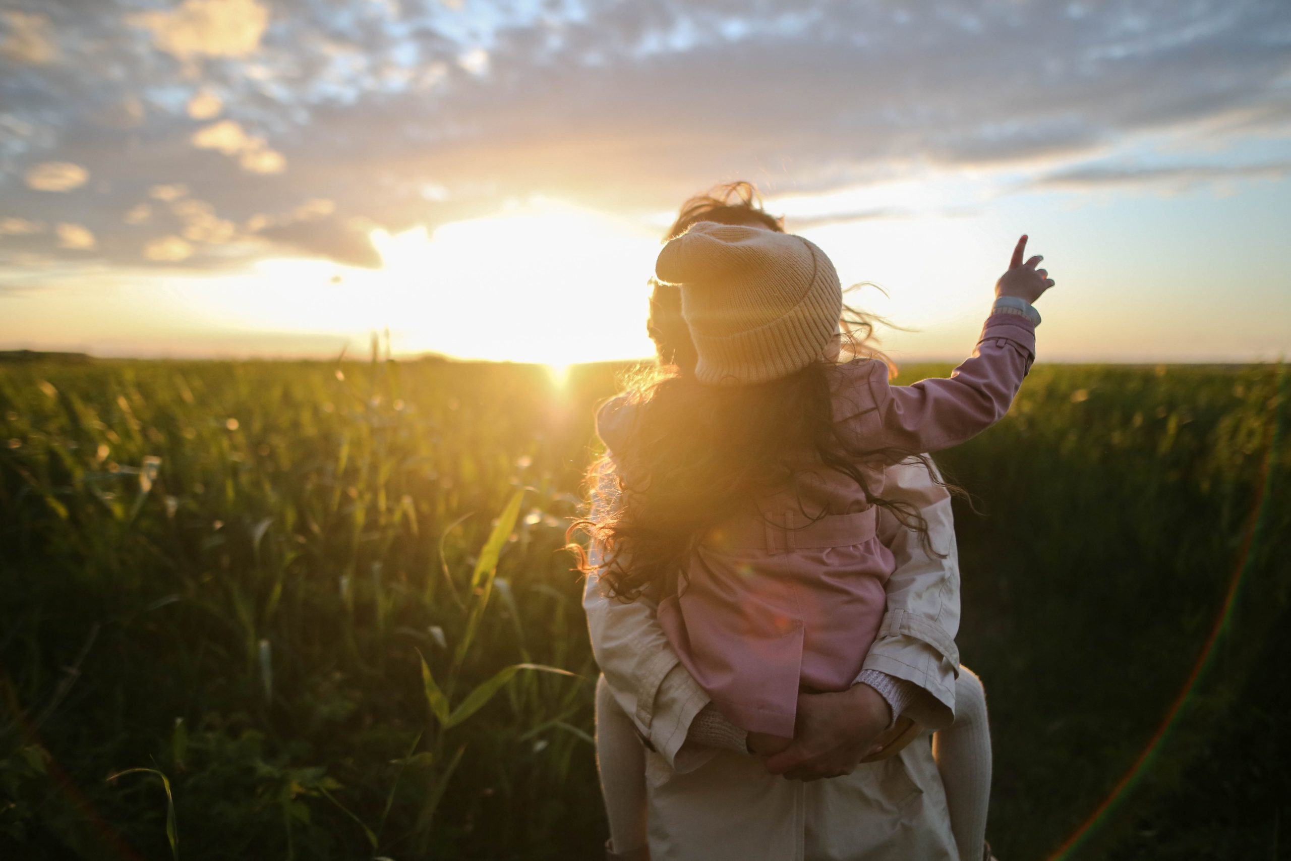 A mother holding her young daughter as they stand in a field, the sun rising behind them.