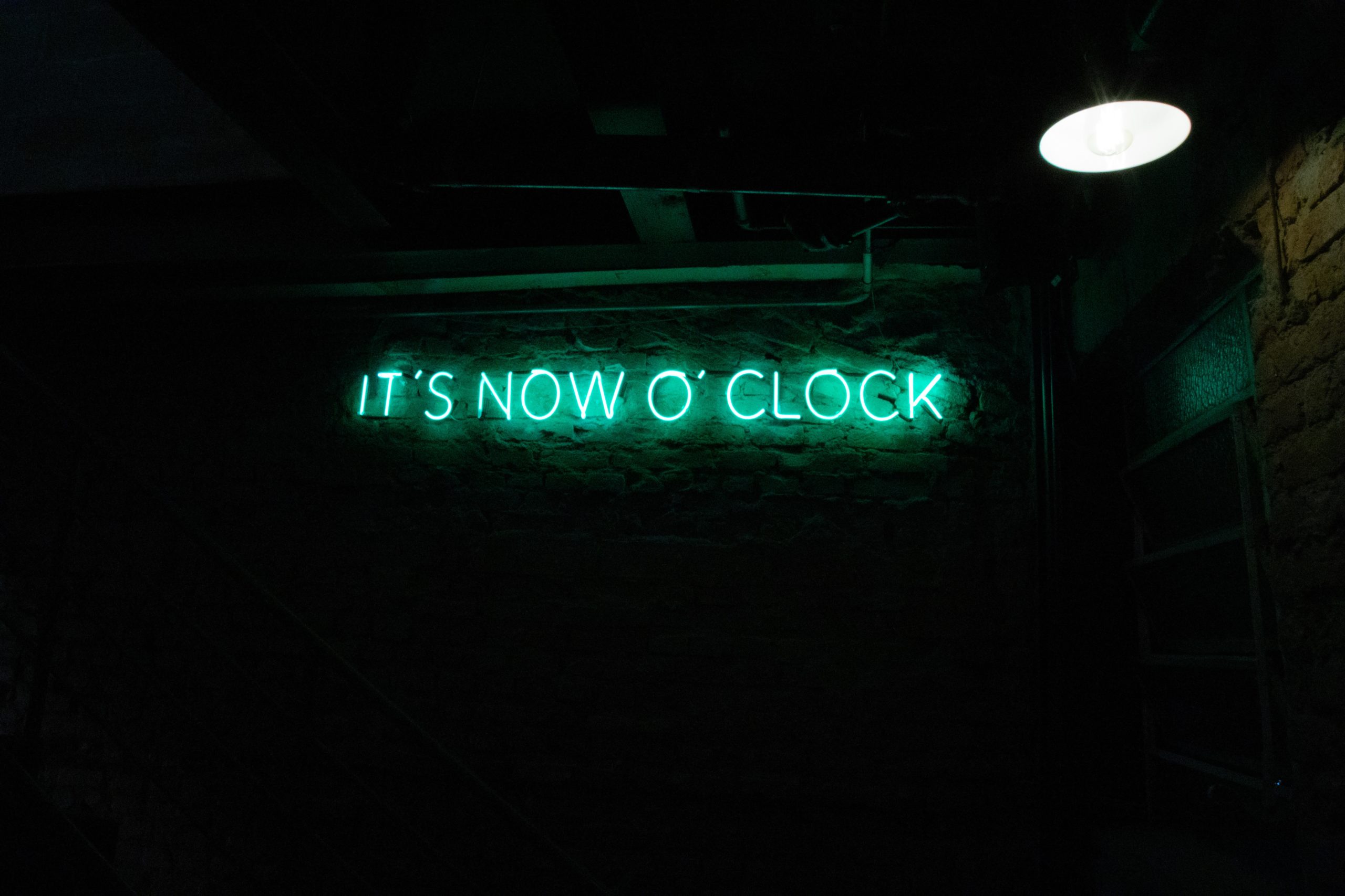 A green neon sign that says 'It's Now O'Clock'.