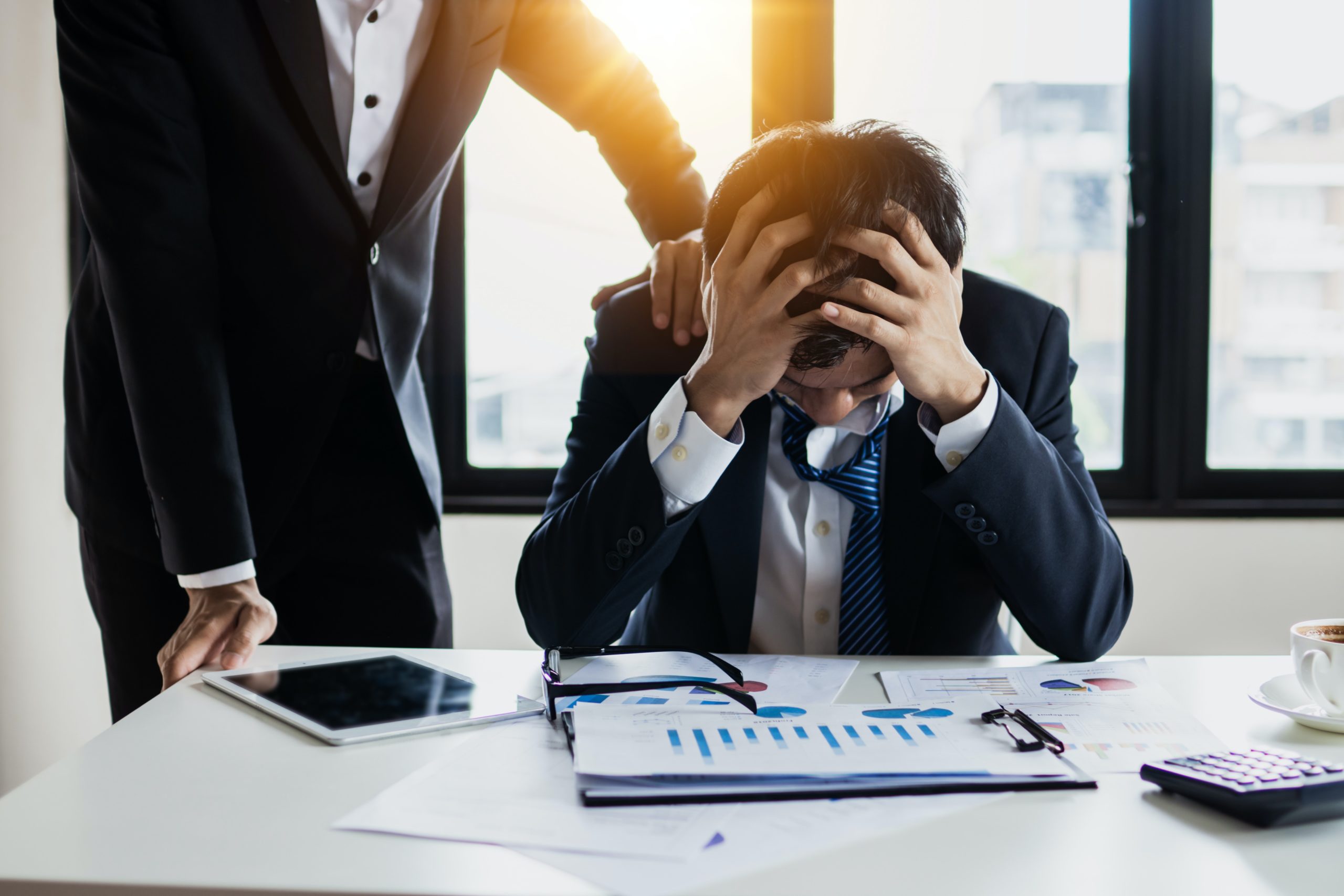 A man with his head in his hands while at work, someone else placing a hand on his shoulder.