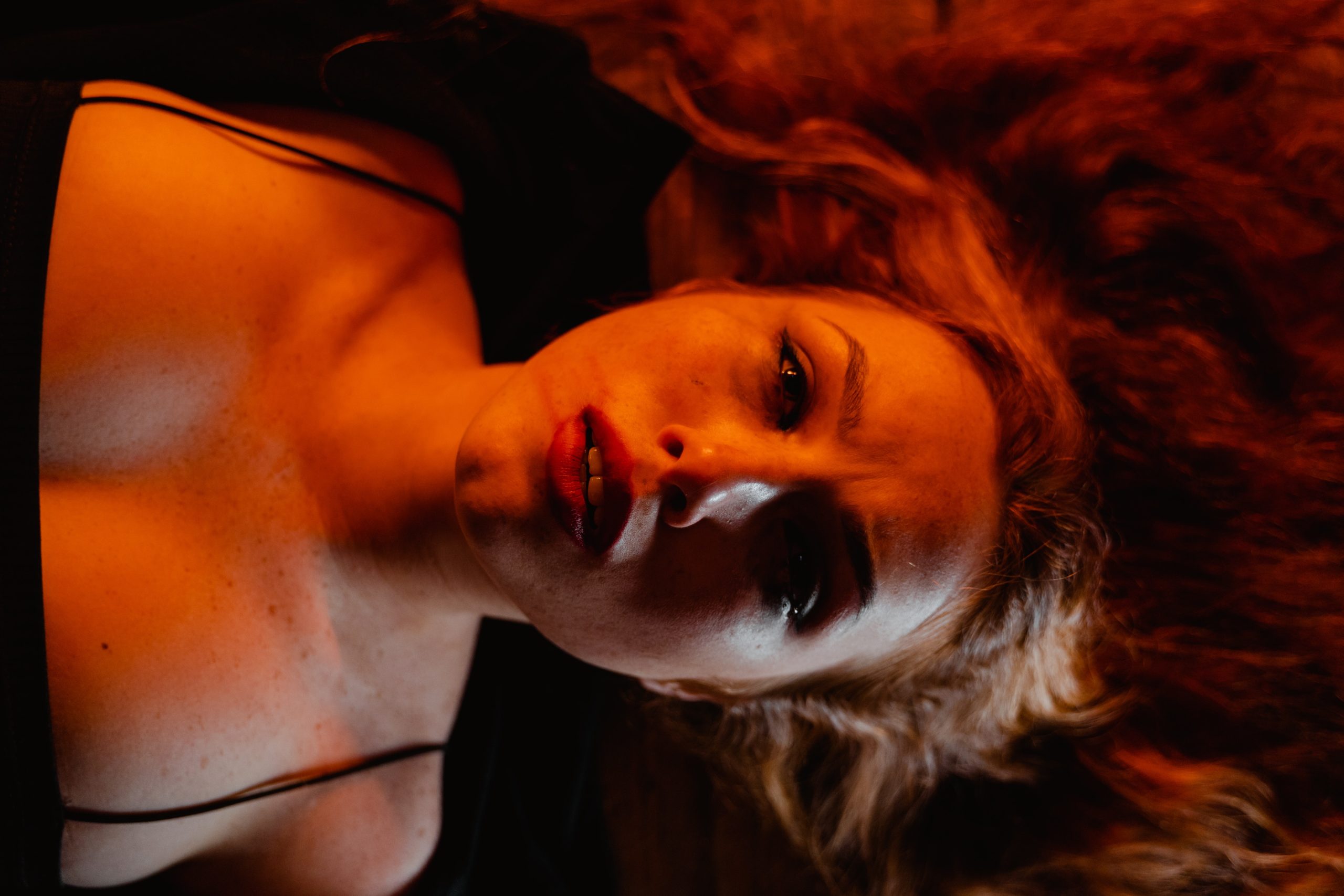 A woman being lit in red, laying down, looking upwards at the camera dramatically.
