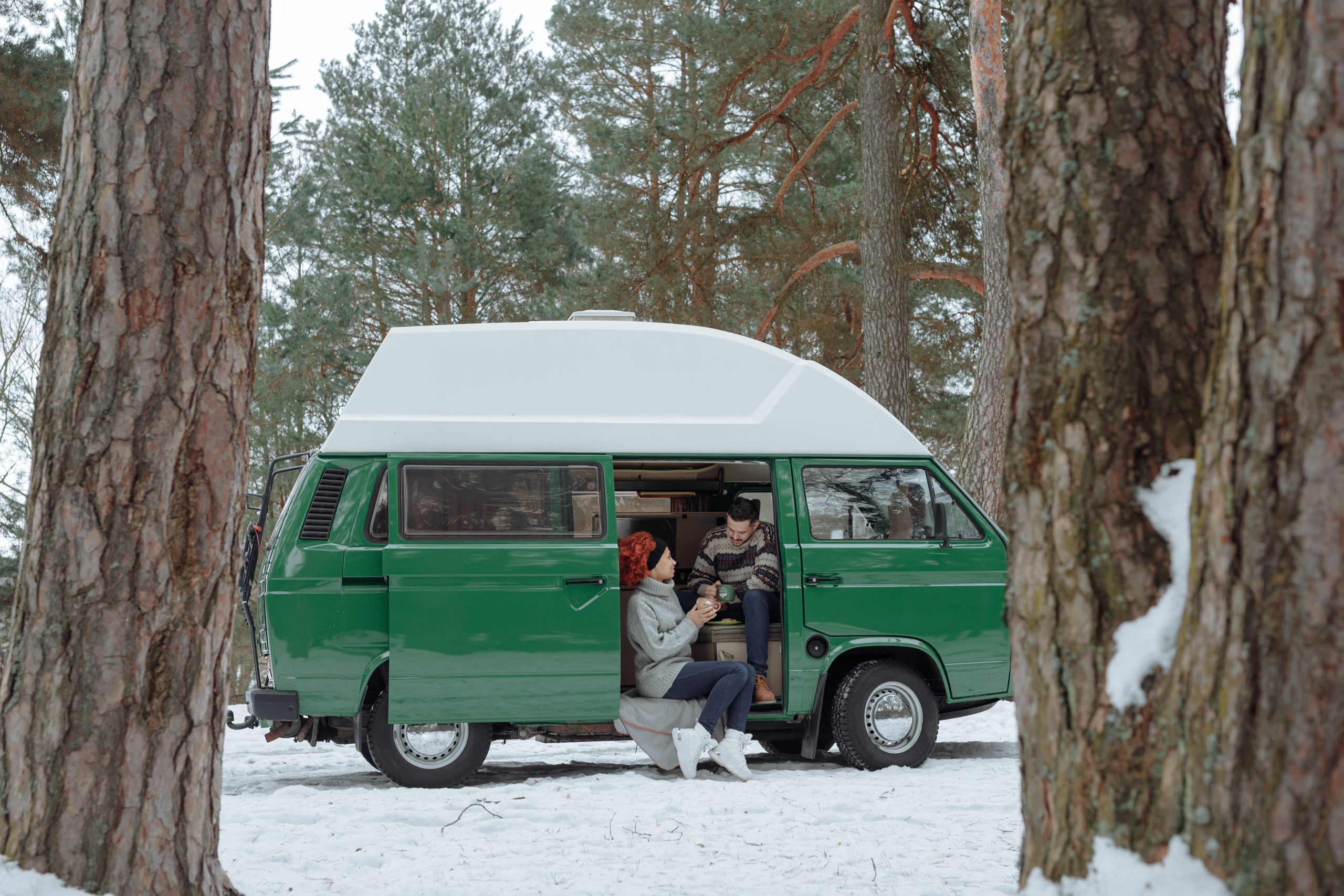 Two people sitting in the door of a van while parked in a snowy forest.