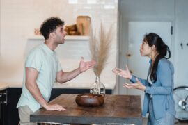 woman in blue denim shirt standing at table arguing with man in white tshirt