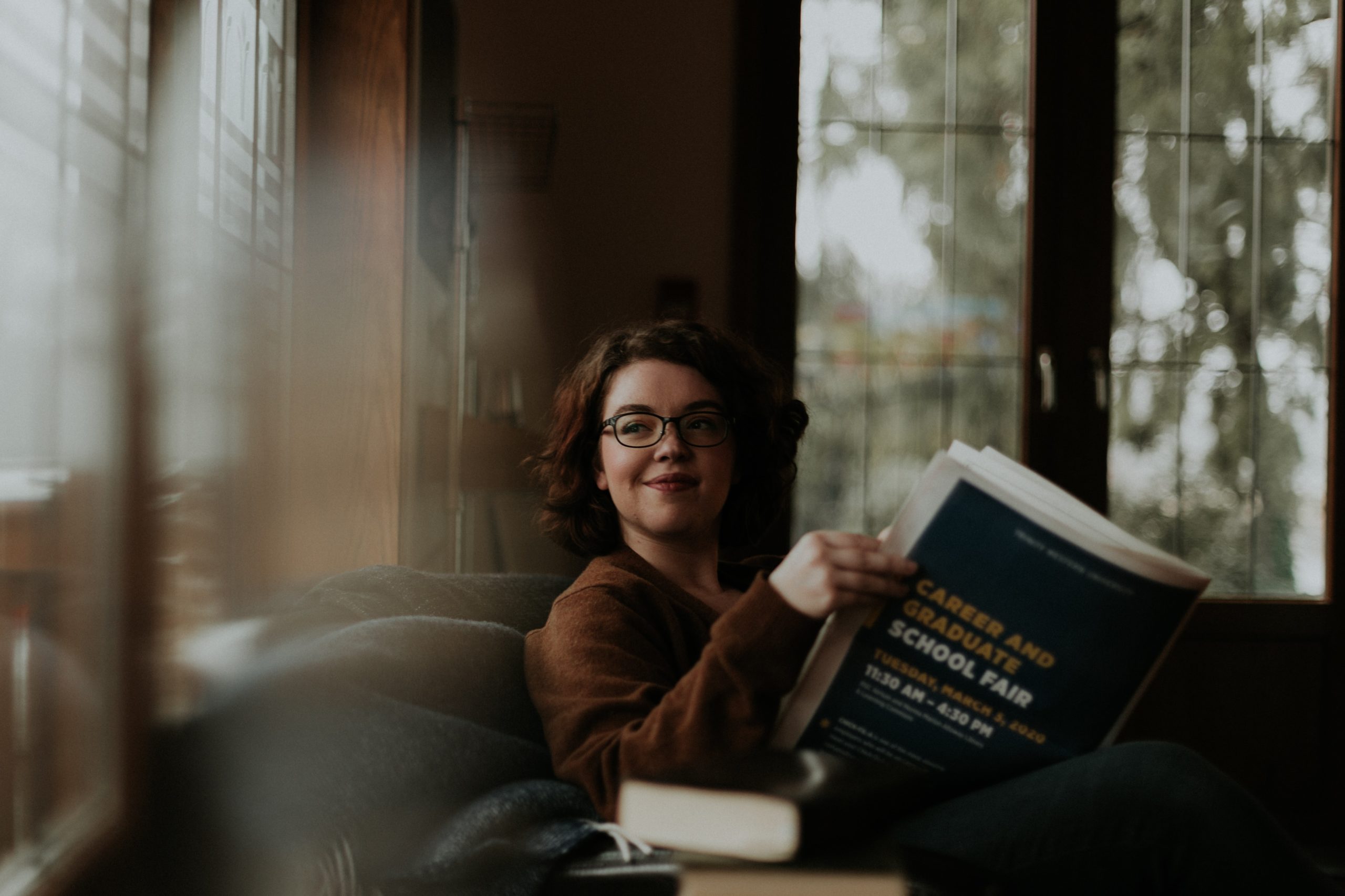 A woman sitting on a couch, reading a newspaper, smiling as she looks out a window.